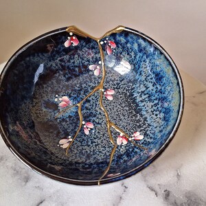 Peut inclure: Un bol en c&eacute;ramique bleu avec une fissure r&eacute;par&eacute;e &agrave; la peinture dor&eacute;e. Le bol est d&eacute;cor&eacute; d'une branche dor&eacute;e et de fleurs roses.