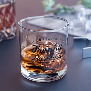 May include: A clear glass with the text "It ain't easy being a Trophy Husband but someone has to" printed on the side. The glass is filled with amber liquid and ice cubes.