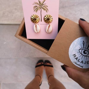 May include: Gold shell earrings with a textured design. The earrings are displayed on a pink card with a palm tree illustration and the text "Let's celebrate".