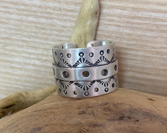 Könnte beinhalten: Ein silberfarbener, verstellbarer Ring mit einem eingravierten geometrischen Muster. Der Ring hat drei gestapelte Bänder mit einem sich wiederholenden Design aus kleinen Kreisen und stilisierten Formen. Der Ring wird auf einem Stück Treibholz präsentiert.