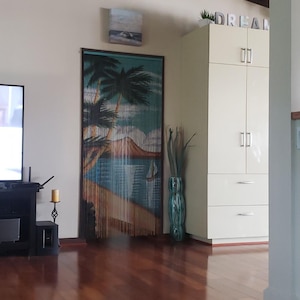 May include: A wooden beaded curtain with a tropical beach scene, featuring palm trees, a blue ocean, and a small island. The curtain is hanging in a doorway with a white cabinet and the word "DREAM" in block letters above it.