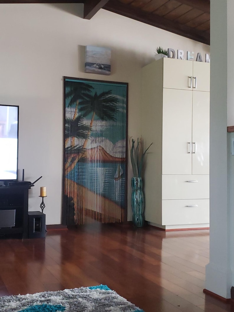 May include: A wooden beaded curtain with a tropical beach scene, featuring palm trees, a blue ocean, and a small island. The curtain is hanging in a doorway with a white cabinet and the word "DREAM" in block letters above it.