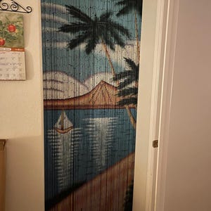 May include: A blue and brown beaded curtain featuring a painted scene of palm trees, a mountain, and a sailboat on a body of water. The beads are strung together to create a decorative room divider.