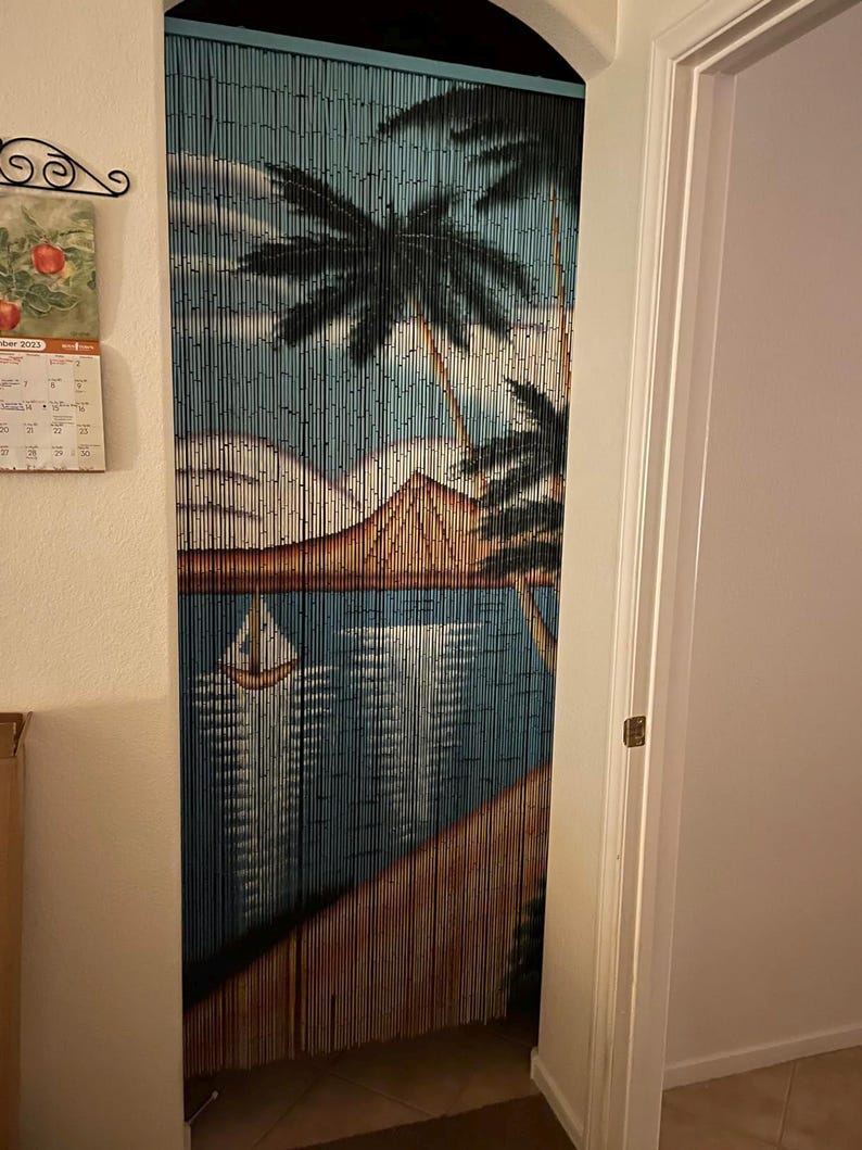 May include: A blue and brown beaded curtain featuring a painted scene of palm trees, a mountain, and a sailboat on a body of water. The beads are strung together to create a decorative room divider.