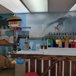 May include: A wooden counter with a variety of colourful syrups in glass bottles. The counter is decorated with a beach scene mural featuring a surfer riding a wave. The mural also includes palm trees and the words "Surf" and "Waikiki".