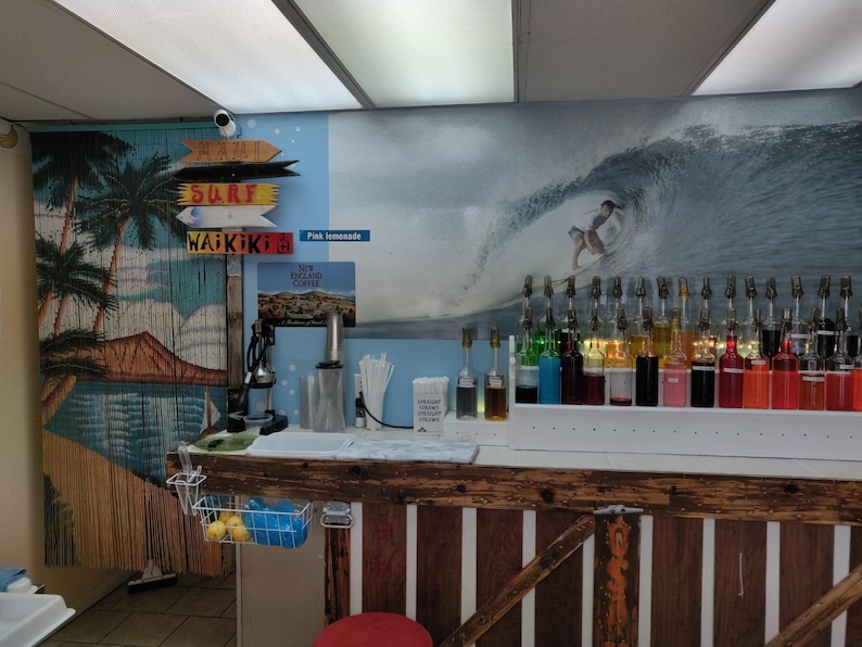 May include: A wooden counter with a variety of colourful syrups in glass bottles. The counter is decorated with a beach scene mural featuring a surfer riding a wave. The mural also includes palm trees and the words "Surf" and "Waikiki".