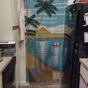 May include: A kitchen with a black refrigerator and a wood-effect floor. A beaded curtain with a tropical beach scene hangs in the doorway. The curtain is made of wooden beads and features palm trees, a beach, and a sailboat.