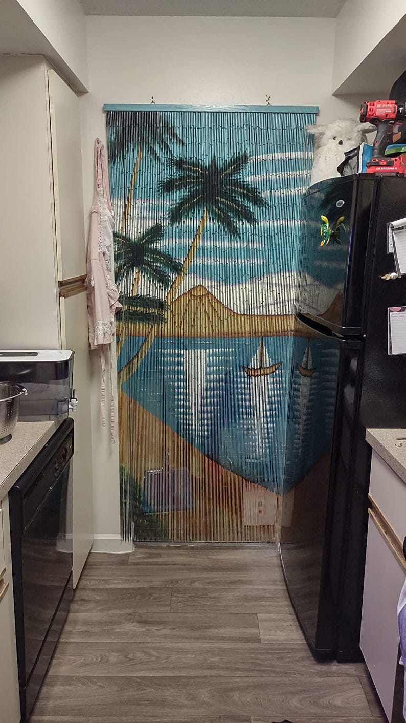 May include: A kitchen with a black refrigerator and a wood-effect floor. A beaded curtain with a tropical beach scene hangs in the doorway. The curtain is made of wooden beads and features palm trees, a beach, and a sailboat.