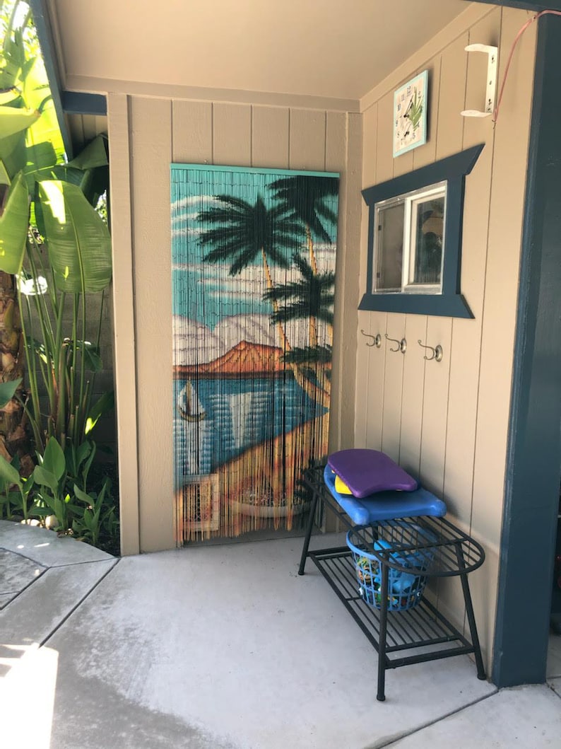 May include: A black metal shelf with a blue basket and a purple and blue surfboard on top. The shelf is in front of a wall with a tropical-themed beaded curtain and a window with a blue frame.