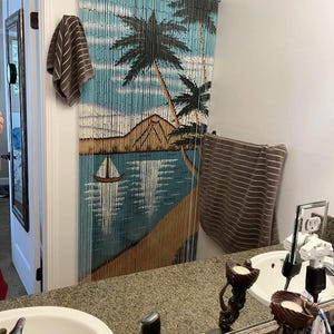 May include: A bathroom with a brown and blue beaded curtain featuring a tropical scene. The curtain depicts palm trees, a beach, and a sailboat. There are two sinks with white porcelain basins and a mirror above each sink. The mirror reflects the bathroom and the beaded curtain.