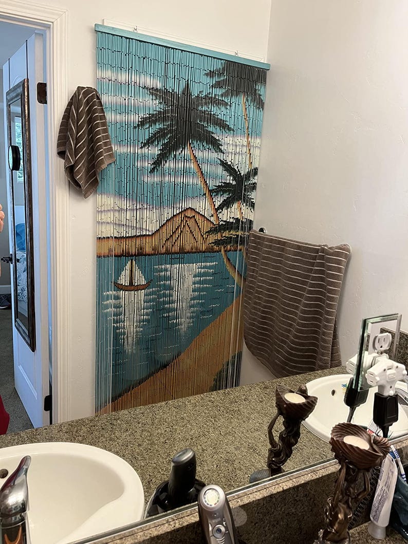 May include: A bathroom with a brown and blue beaded curtain featuring a tropical scene. The curtain depicts palm trees, a beach, and a sailboat. There are two sinks with white porcelain basins and a mirror above each sink. The mirror reflects the bathroom and the beaded curtain.