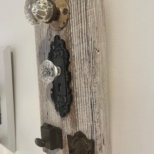 Puede incluir: Un perchero de pared hecho de una pieza rectangular de madera blanca desgastada. Cuenta con un pomo de cristal transparente, una placa de metal negro con un pomo de cristal, un pequeño pestillo de metal oscuro y un tirador de cajón de latón.