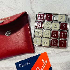 May include: A square sliding tile puzzle with alternating dark red and cream-colored numbered squares, accompanied by a red storage pouch with a silver snap button and a blue and orange card labeled Known the Puzzle.