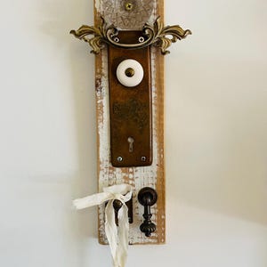 Könnte beinhalten: Ein rustikaler Wandbehang mit Vintage-Hardware auf einer verwitterten Holzplanke. Er enthält einen durchsichtigen Glas-Türknauf, Messingakzente, einen weißen Keramikknauf und einen dekorativen Schlüssel. Das Holz ist weiß gestrichen und hat ein verwittertes Finish.