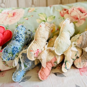 May include: A collection of handmade fabric bunny decorations in various floral patterns and colors, including red, blue, and cream. Each bunny is tied with a white ribbon. The bunnies are arranged on a floral patterned pillow.