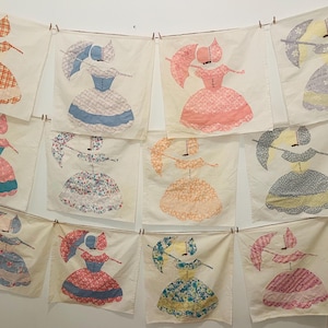 May include: Twelve framed fabric squares with embroidered images of women wearing dresses and holding umbrellas. The squares are hung on a white wall with clothespins.