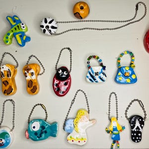 May include: A collection of 14 handmade ceramic ornaments, including a ladybug, a fish, a butterfly, a snowman, a girl, an angel, a pair of flip-flops, and several purses. The ornaments are all brightly colored and feature intricate details.