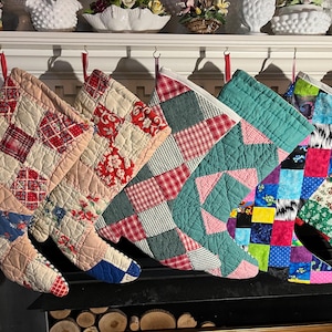 May include: Six quilted Christmas stockings hanging in a row. The stockings feature various patchwork designs including floral, plaid, and geometric patterns in colors such as red, green, blue, yellow, and cream with stitched textures.