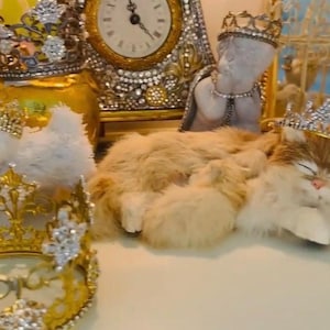 May include: A collection of decorative items including a clock, figurines, and crowns. A white dog and a cat are wearing jeweled crowns. The scene is set with a vintage aesthetic, featuring gold and crystal accents.