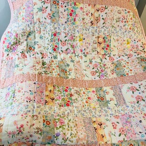 May include: A pink and white floral patchwork quilt with a pink border. The quilt is made of various floral fabrics in shades of pink, white, and green.