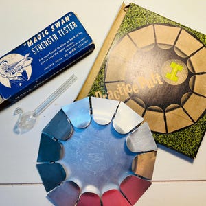 May include: An assortment of vintage items, including a blue box labeled "MAGIC SWAN STRENGTH TESTER" with a swan-shaped glass whistle, a "Practice Putt" game, and a multi-colored metal object.