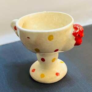 May include: A cream-colored ceramic egg cup featuring a hand-painted red rooster head on one side and a tail-like handle on the other. The exterior is decorated with small yellow and red polka dots, and the surface has a visible crackle glaze finish.