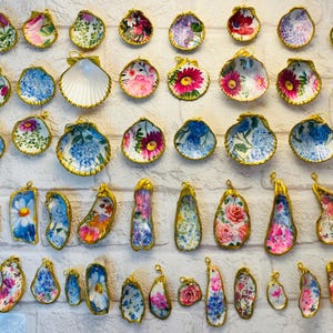 May include: A collection of assorted seashells featuring various floral and botanical patterns in vibrant colors. Each shell is finished with a metallic gold painted edge and includes a small gold loop at the top for hanging as a decorative ornament.