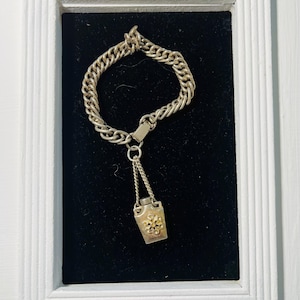 May include: A silver-toned bracelet with a chain link design, featuring a small rectangular charm with a floral design. The bracelet is displayed on a black background within a white framed shadow box.