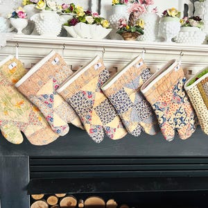 May include: Six quilted oven mitts hanging in a row, each featuring a unique patchwork design with floral, geometric, and polka dot patterns in shades of yellow, blue, red, and brown. Each mitt has a white cuff and a small numbered tag.