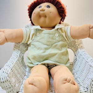 May include: A vintage Cabbage Patch Kid doll with red yarn hair, wearing a light green corduroy outfit. The doll is seated in a white wicker chair, arms outstretched.