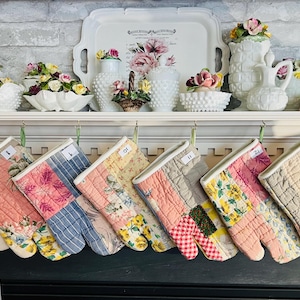 May include: A row of quilted fabric oven mitts hanging from a mantel, each featuring a unique patchwork design with floral, plaid, and gingham patterns in soft pastel colors. Each mitt is numbered with a small white tag attached to the top edge.