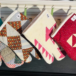 May include: Four quilted oven mitts hanging in a row, numbered one through four. The mitts feature various patchwork designs in red, cream, pink, teal, and multi-colored floral patterns, each with a white fabric cuff and a green hanging loop.