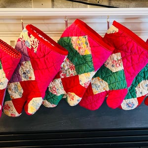 May include: A row of quilted oven mitts in various colors, including red, pink, green, and purple. The mitts are hanging from a white shelf. The mitts have a patchwork design with floral patterns.