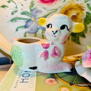 May include: A ceramic lamb planter with a white body, pink accents, and yellow ears. The lamb has a smiling face and is decorated with pink bows. The planter is on a book with the words "HOME COMPANY" and a floral tray in the background.