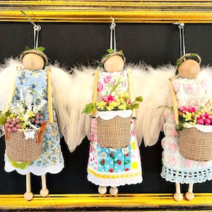 May include: Three handmade angel ornaments with wooden heads, feather wings, and fabric dresses. Each angel carries a burlap basket filled with flowers and greenery. The angels are suspended within a gold framed black background.