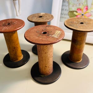 May include: Four vintage wooden spools with a dark brown base, a light brown cylindrical body, and a reddish-brown top. The spools are arranged on a light-colored surface, with a framed picture in the background.