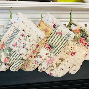 May include: Three quilted oven mitts with floral and striped patterns. Each mitt has a different design, including roses and stripes in shades of pink, yellow, and green. Numbers 1, 2, and 3 are visible on the top of each mitt.