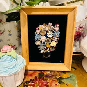 May include: A rectangular wooden frame containing a black velvet backing adorned with a cluster of assorted vintage-style jewellery pieces, including small floral-shaped brooches, faux pearls, rhinestones, and a single clear teardrop-shaped crystal.