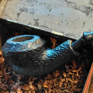 May include: A black tobacco pipe with a textured bowl and a curved, glossy black stem. The pipe rests inside a metal tin filled with loose tobacco. The background is a wooden surface.