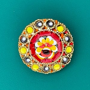 May include: A round mosaic brooch with a gold-tone metal frame. The mosaic features a central floral design in yellow, red, and white, surrounded by red and yellow tiles, and black and white accents.