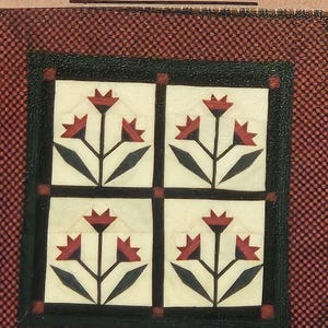 Könnte beinhalten: Ein dekoratives Quadrat mit vier kleineren Quadraten, die jeweils ein stilisiertes Lilienmuster zeigen. Die Lilien haben rote Blütenblätter, schwarze Stiele und Blätter, vor einem cremefarbenen Hintergrund, umrahmt von einem dunkelgrünen Rand. Das Wort "Lilies" steht oben.