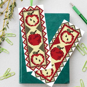 May include: Two handmade bookmarks with a cross-stitch apple design. The bookmarks feature a red apple on a cream background, framed by a red and brown border. One bookmark is placed on a green book, with a pen and paperclips nearby.