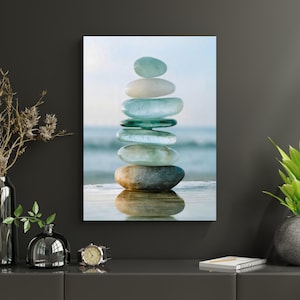 May include: A vertical wall art print featuring a stack of seven smooth, rounded stones in shades of pale blue, white, and grey. The stones are balanced on a reflective surface with a blurred ocean and sky background, creating a serene composition.