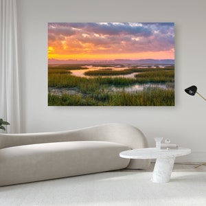 May include: A photograph of a marsh landscape at sunrise. The sky is a vibrant mix of pink, orange, and purple hues. The marsh is covered in green grasses and water, with a few small islands scattered throughout.