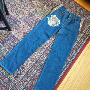May include: Blue denim jeans with a colorful embroidered patch of a leopard on the front left leg. The jeans have a yellow and blue patterned fabric trim along the sides.