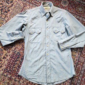 May include: A light blue long-sleeved button-down shirt with a white stripe pattern. The shirt has two chest pockets with a pointed flap and a single button closure.