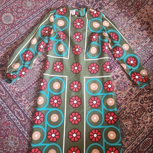May include: Olive green dress with a floral pattern in red, tan, and teal. The dress has long sleeves and a knee-length hemline. The floral design is arranged in a grid-like pattern with white outlines.