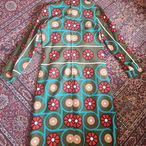 May include: Olive green dress with a vibrant geometric pattern. The design features red flower-like shapes, teal circles, and tan and white accents. The long-sleeved dress is laid flat, showcasing its retro style.