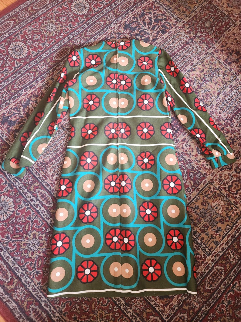 May include: Olive green dress with a vibrant geometric pattern. The design features red flower-like shapes, teal circles, and tan and white accents. The long-sleeved dress is laid flat, showcasing its retro style.