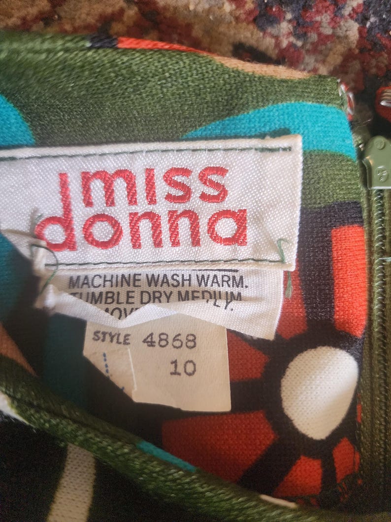 May include: Close-up of a vintage dress label with the text "miss donna" in red. The label also includes washing instructions and a size 10 tag. The dress features a colorful floral pattern with green, blue, red, and white elements.
